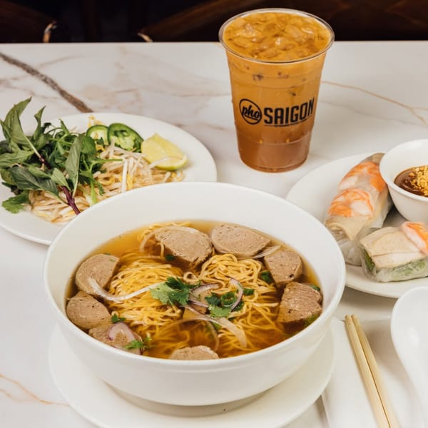 Noodle soup with meatballs, fresh herbs, spring rolls, and an iced drink from Pho Saigon on a marble table.