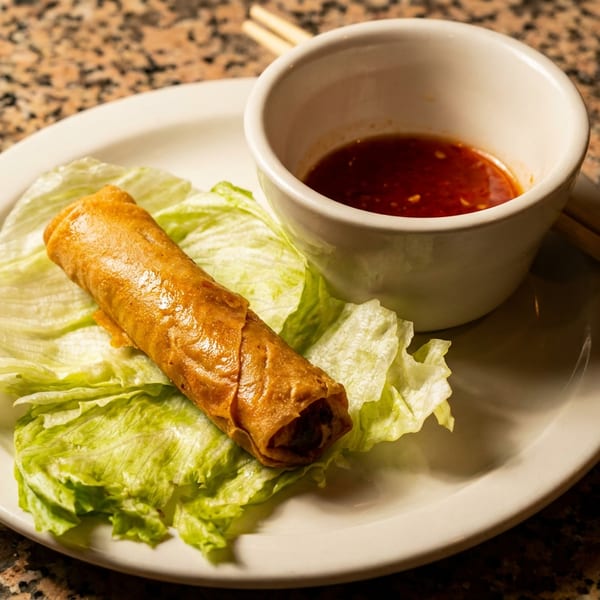 Golden-brown spring roll on lettuce with red dipping sauce and chopsticks on a white plate.