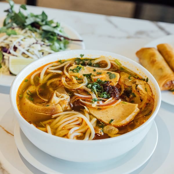 Spicy Vietnamese noodle soup with various meats, herbs, and spring rolls on a white marble table.